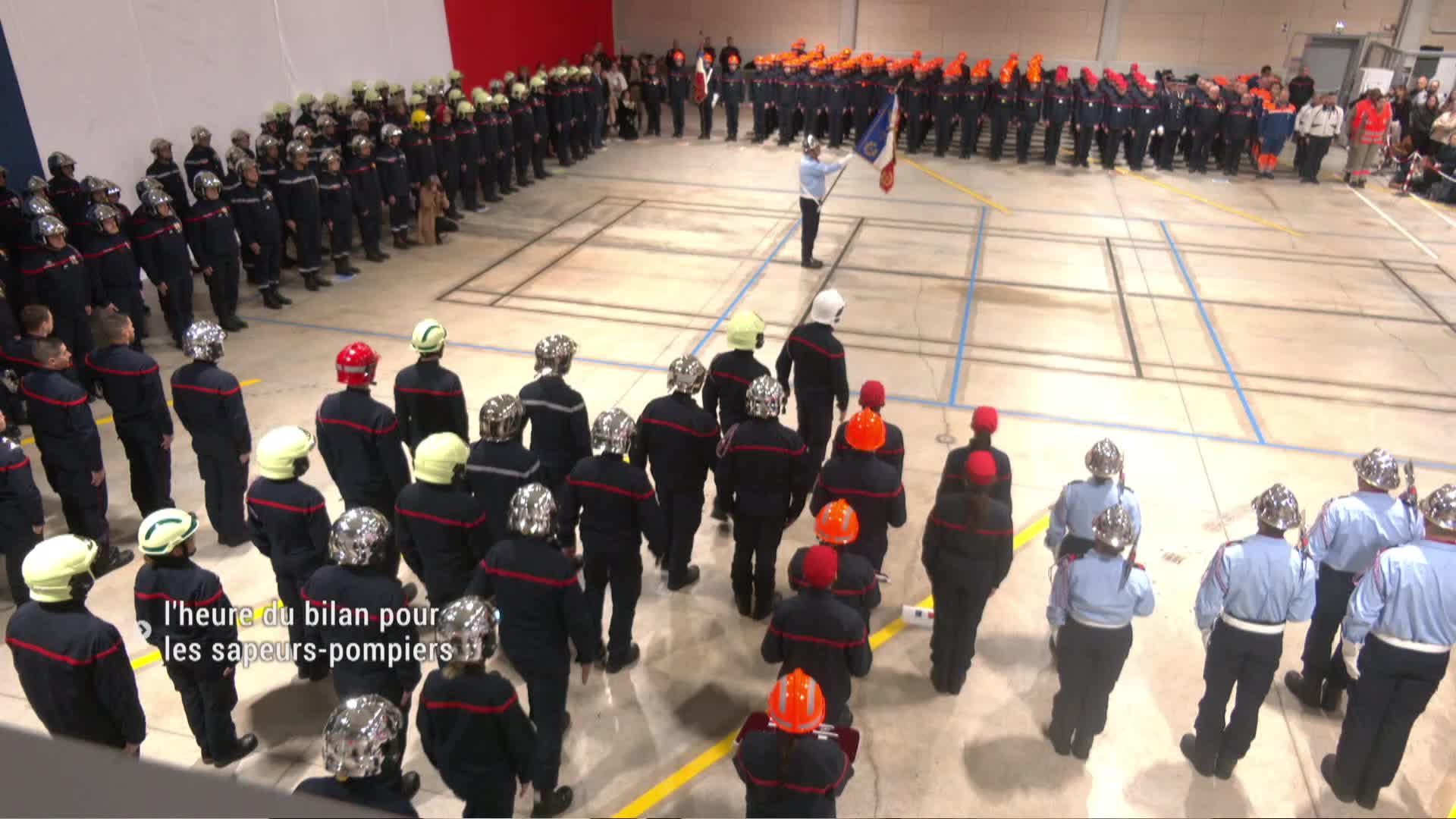 Pompiers en uniforme noir et rouge rassemblés dans un gymnase lors d'une cérémonie officielle.
