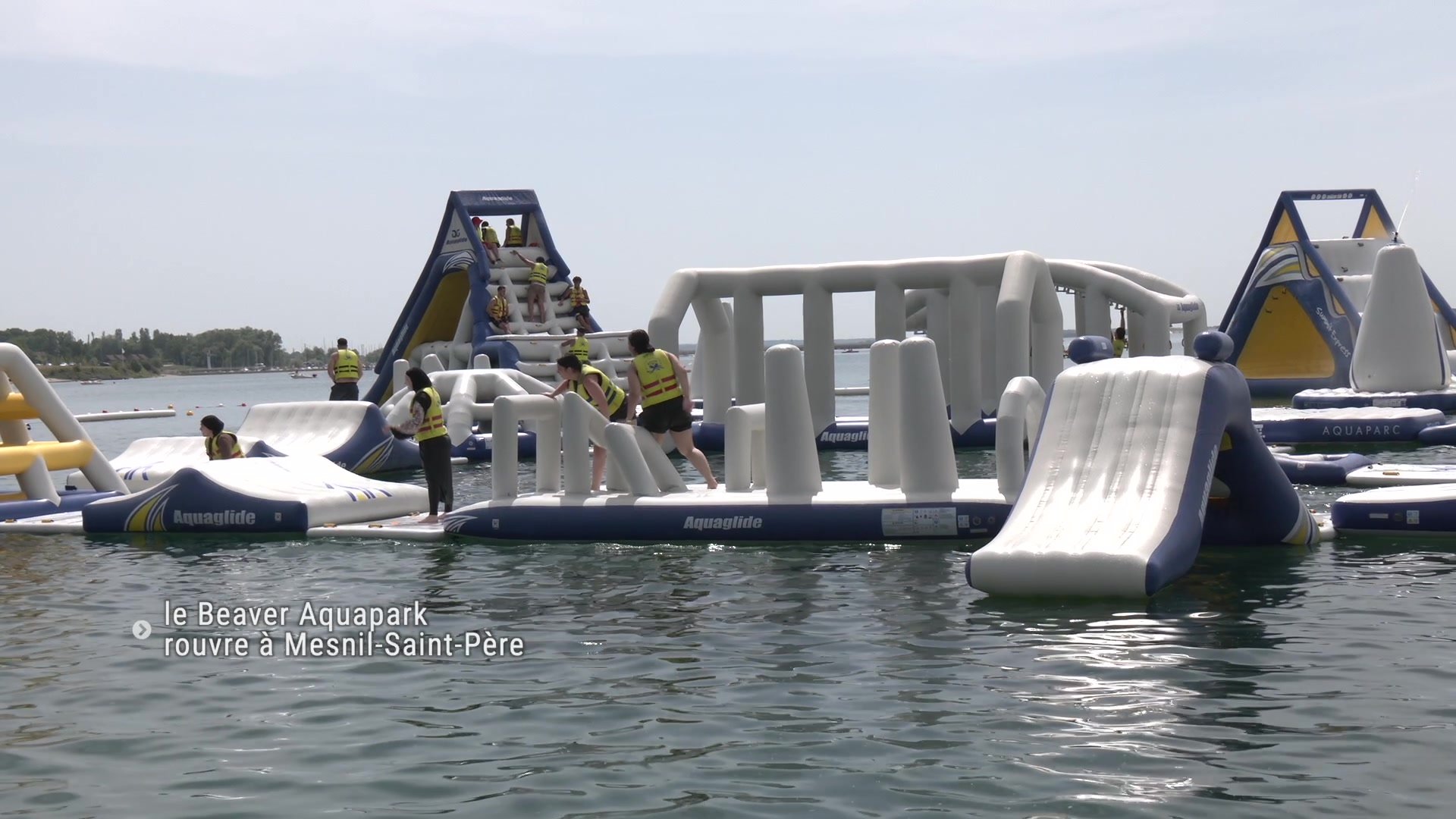Structure gonflable du Beaver Aquapark avec des baigneurs en gilets jaunes sur des modules bleus et blancs posés sur un lac