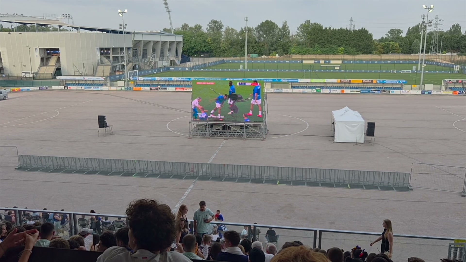 Écran géant diffusant un match de football dans un stade presque vide, spectateurs en tribune au premier plan