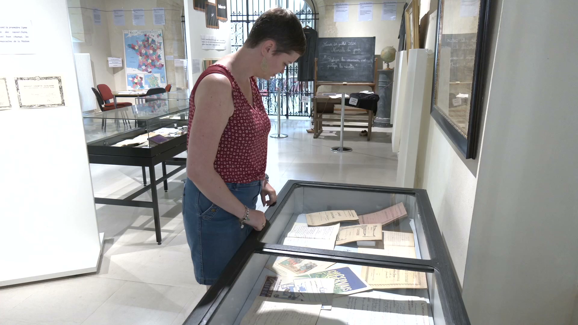 Femme consultant des cahiers d'écoliers anciens dans une vitrine au musée Napoléon, tableau noir et globe terrestre visibles en arrière-plan