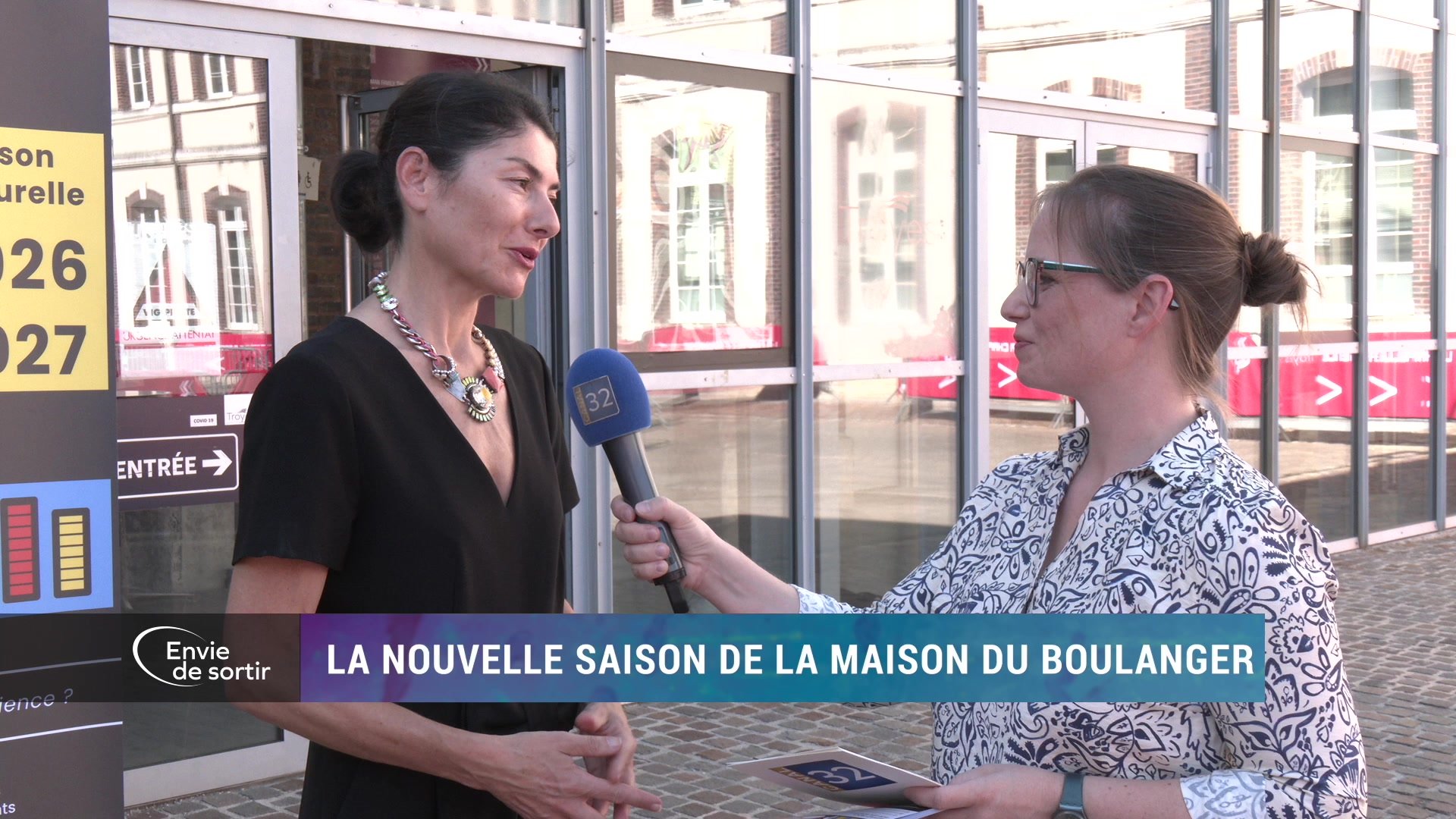 Journaliste tendant un micro Canal 32 à une femme devant l'entrée de La Maison du Boulanger