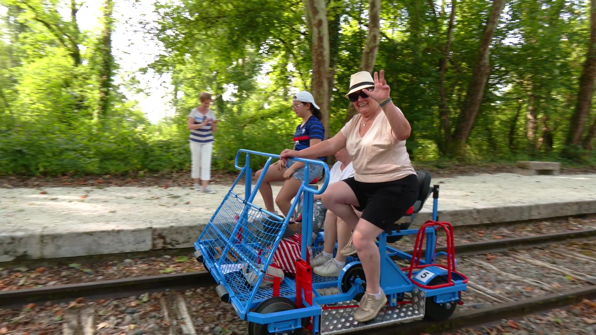 Participantes à bord d'un vélo-rail bleu sur les rails en forêt, l'une d'elles salue la caméra avec un chapeau de paille