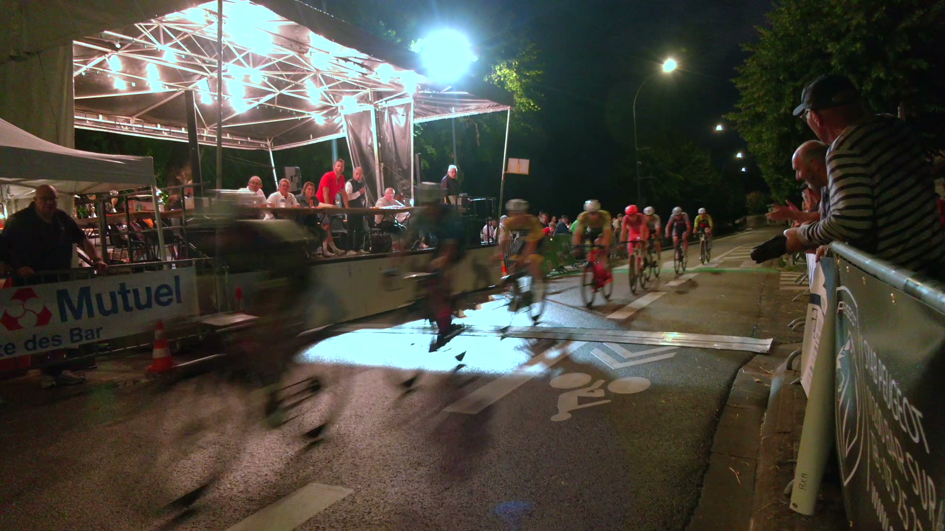 Peloton de cyclistes en pleine vitesse la nuit sur circuit éclairé, spectateurs derrière barrières métalliques
