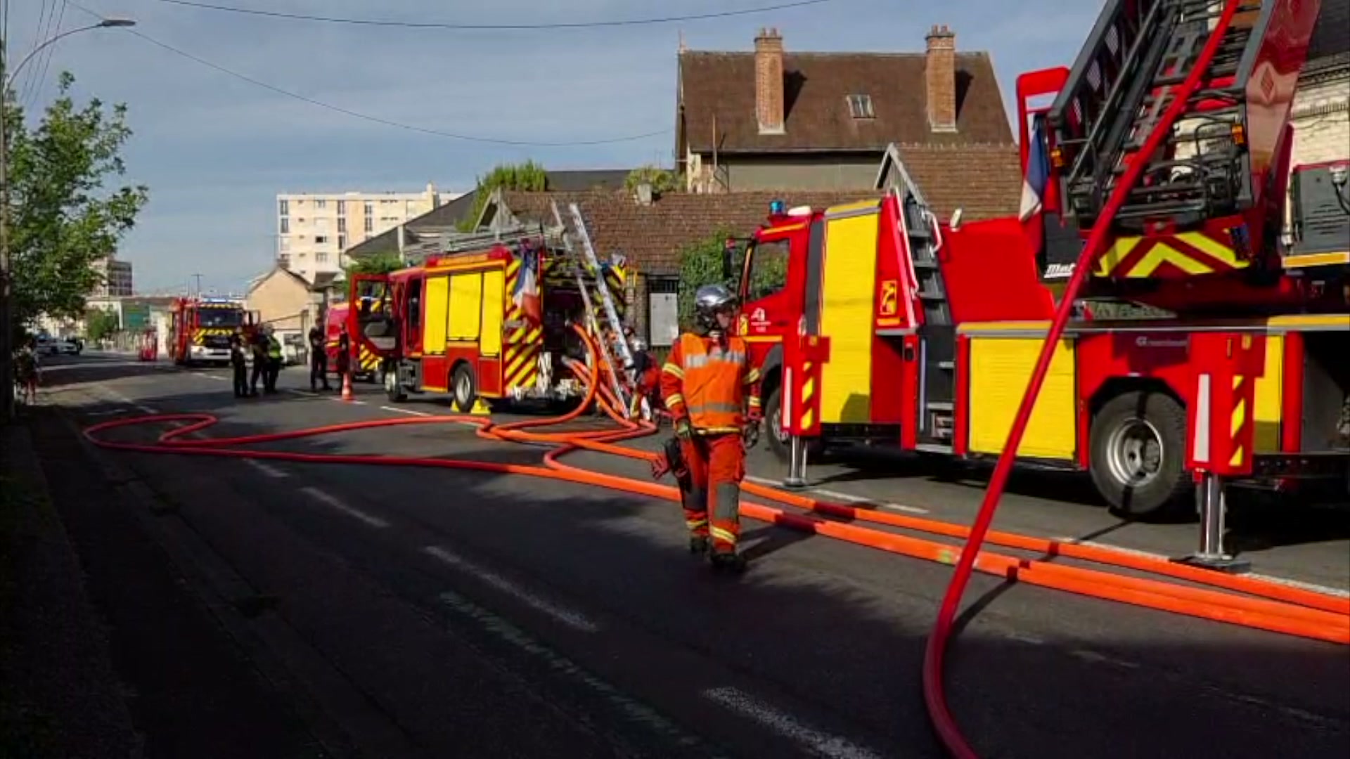 Pompier en tenue orange marchant sur la chaussée entre des lances à incendie déployées, camions rouges et grande