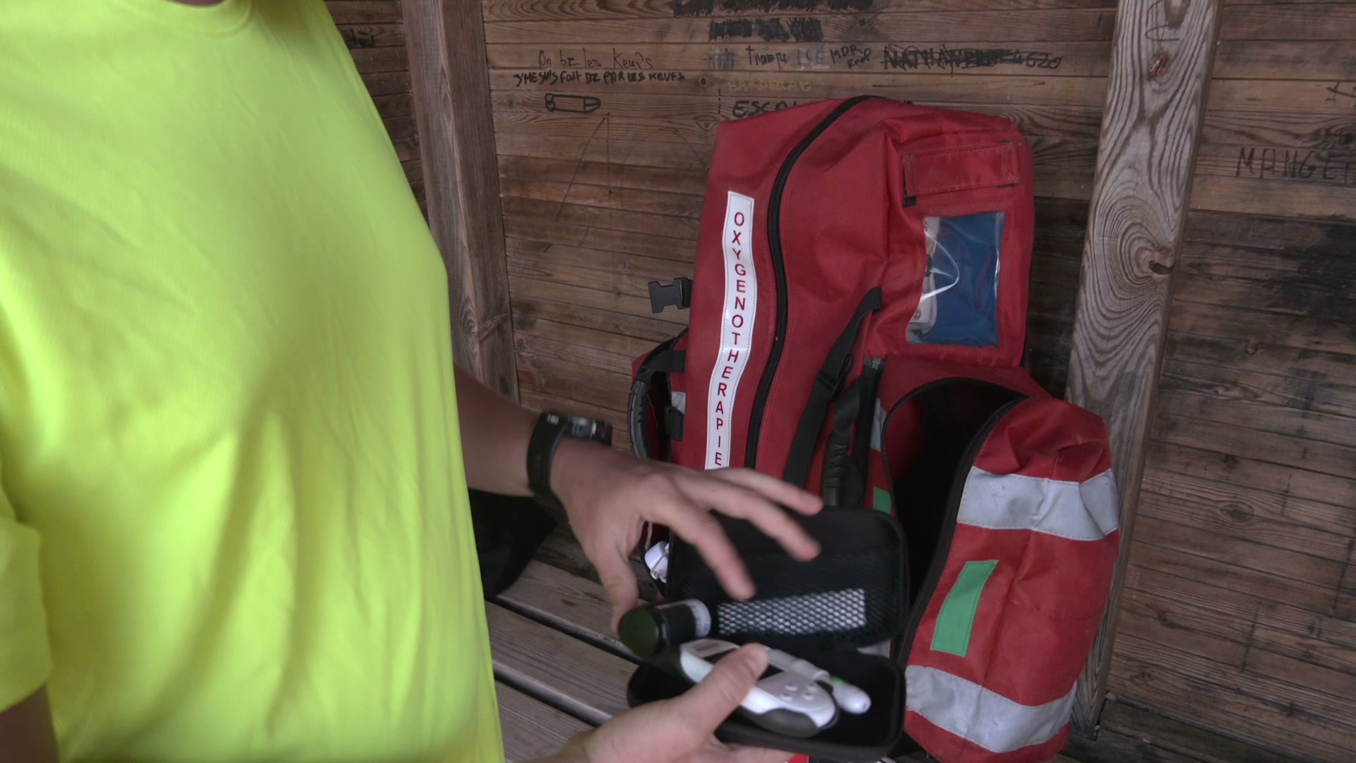 Surveillant en t-shirt jaune fluo vérifiant un appareil de mesure devant un sac rouge d'oxygénothérapie