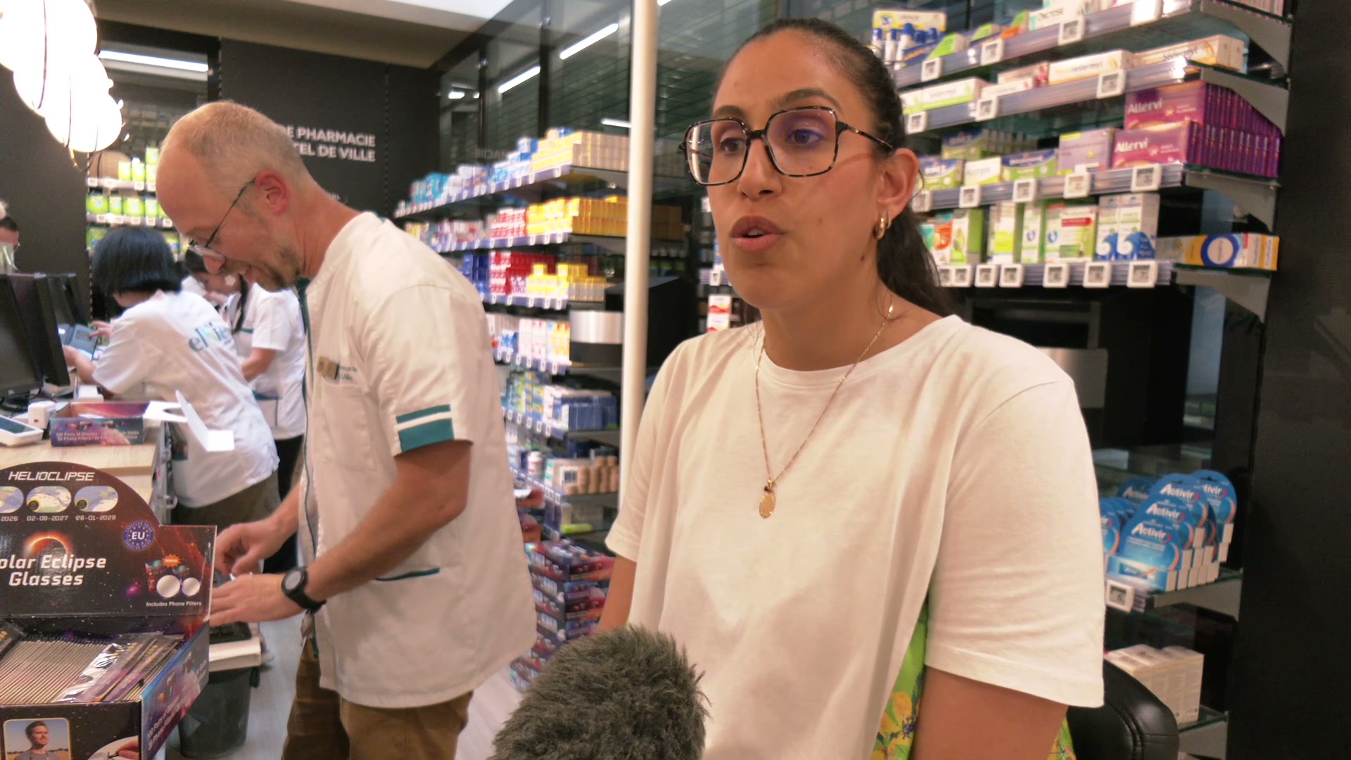 Cliente interviewée devant un présentoir de lunettes Solar Eclipse Glasses dans une pharmacie de Troyes