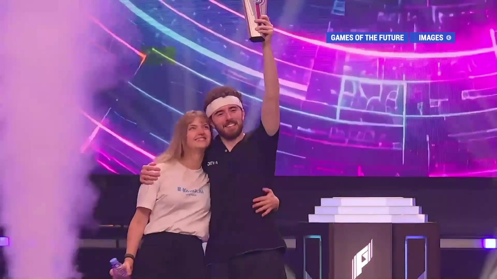 Deux compétiteurs sur scène aux Games of the Future, l'un brandissant un trophée devant un écran LED violet et rose