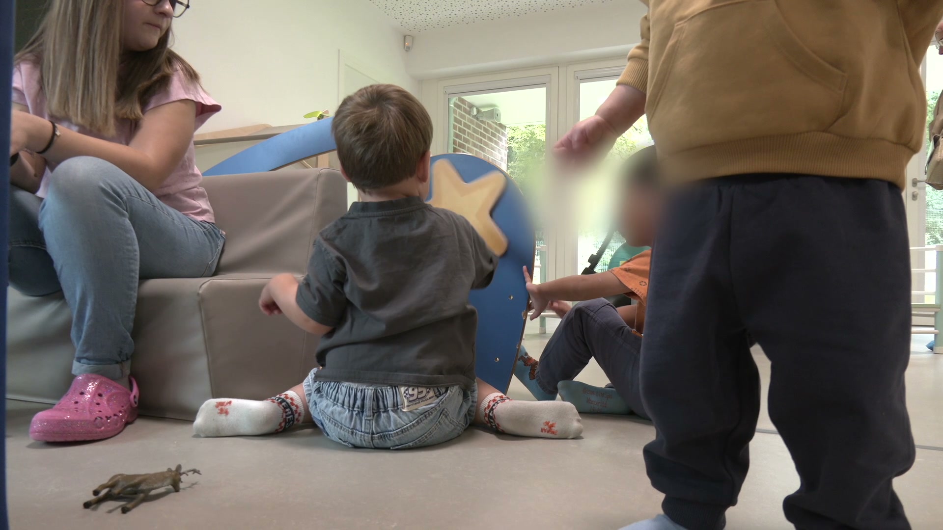 Enfants assis et debout sur le sol d'une salle de jeux lumineuse, avec un module en mousse bleu étoilé et une auxiliaire en crocs roses