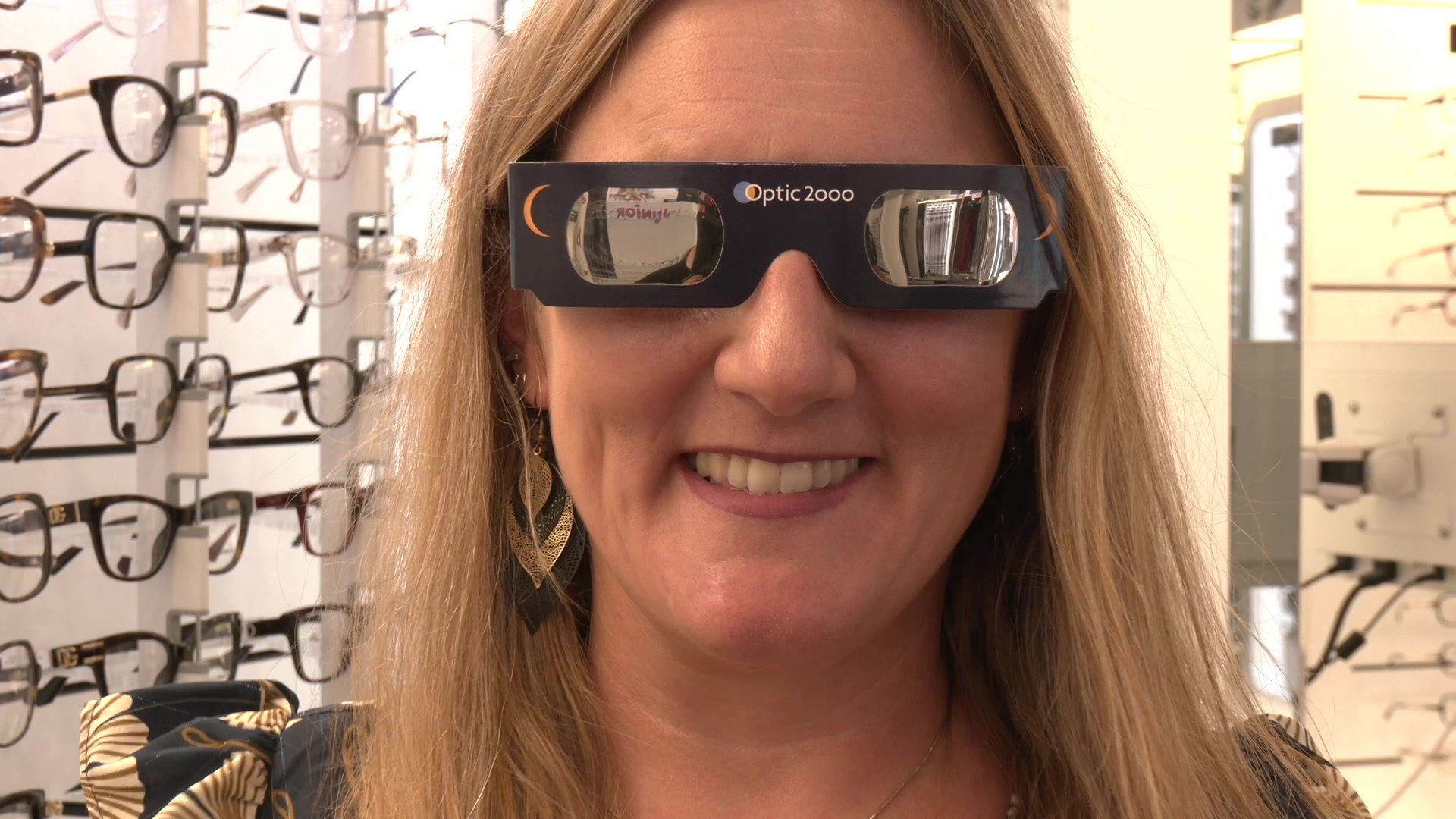 Femme souriante portant des lunettes éclipse Optic 2000 devant un présentoir de montures