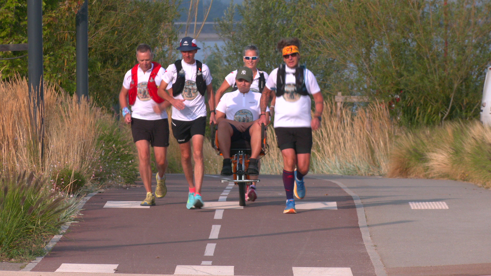 Quatre coureurs en tee-shirt blanc poussent une joëlette sur une piste cyclable bordée de végétation au lac d'Orient