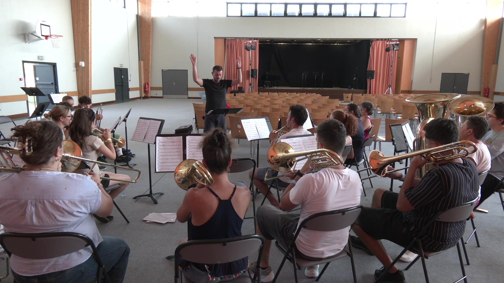Stagiaires musiciens en répétition encadrés par un chef les bras levés dans une salle omnisports