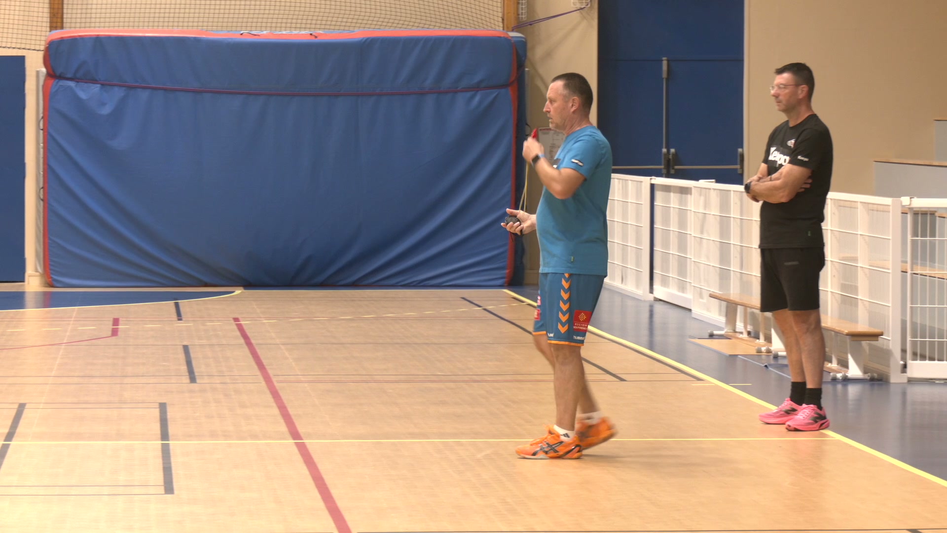 Deux entraîneurs debout sur un parquet de salle de handball, l'un en tenue bleue hummel tenant un sifflet