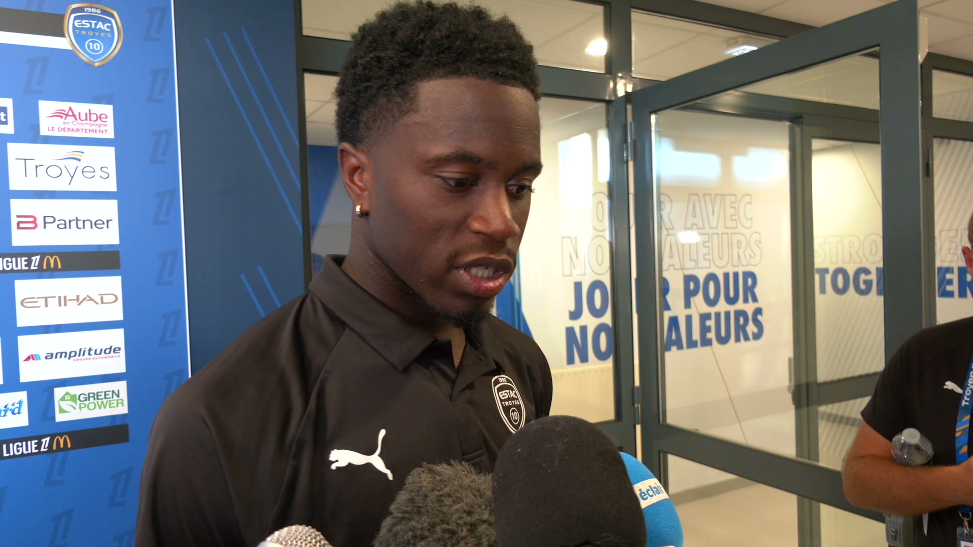 Junior Diaz en polo noir Estac Troyes face à des micros, devant un panneau sponsors au Stade de l'Aube