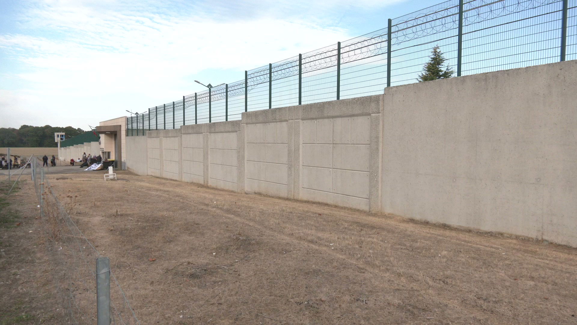 Mur de béton surmonté de grillages barbelés verts du centre de détention, agents rassemblés près de l'entrée avec une banderole