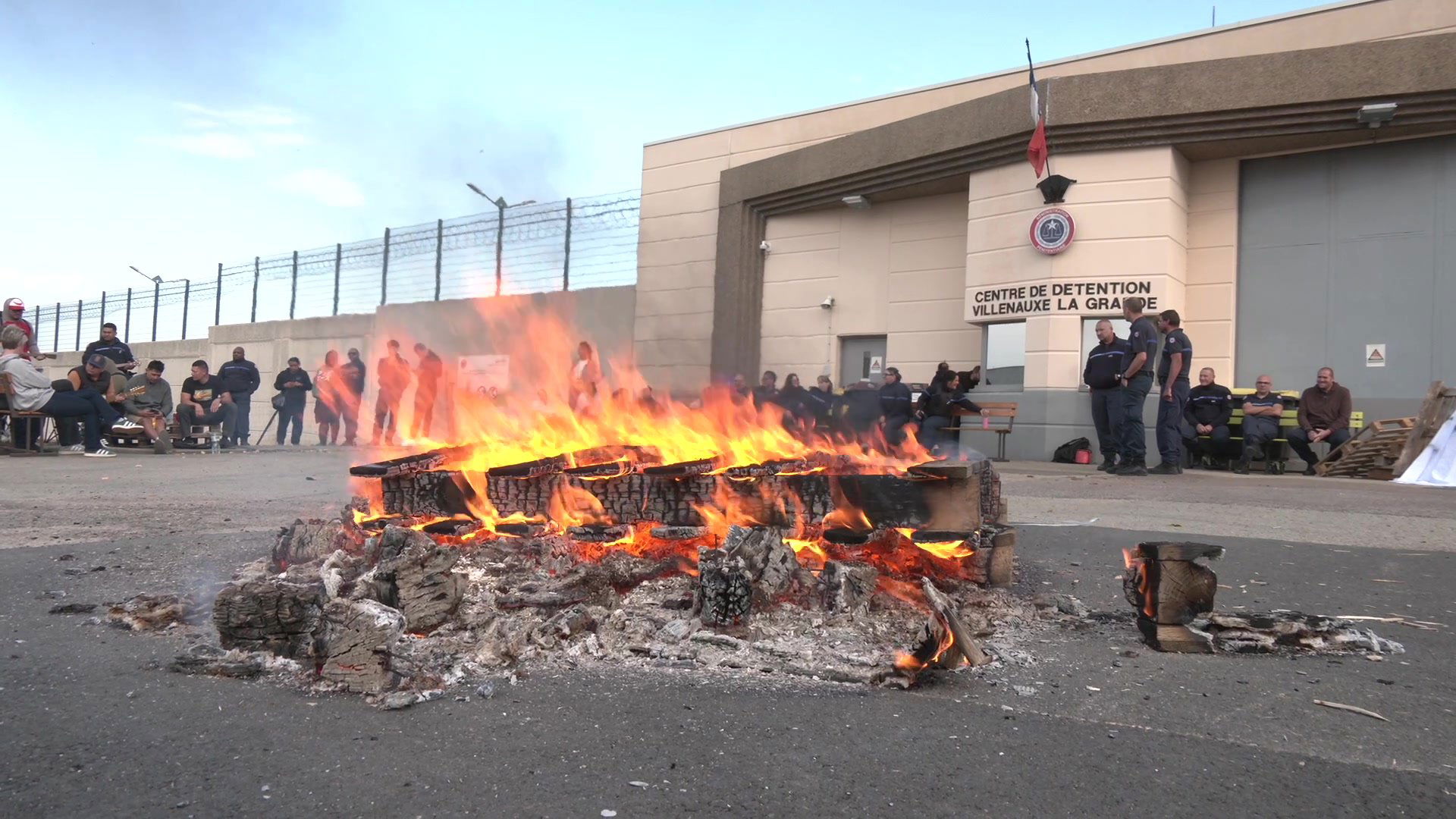 Mur d'enceinte en béton surmonté de grillages barbelés du centre de détention de Villenauxe-la-Grande, avec des agents rassemblés devant l'entrée