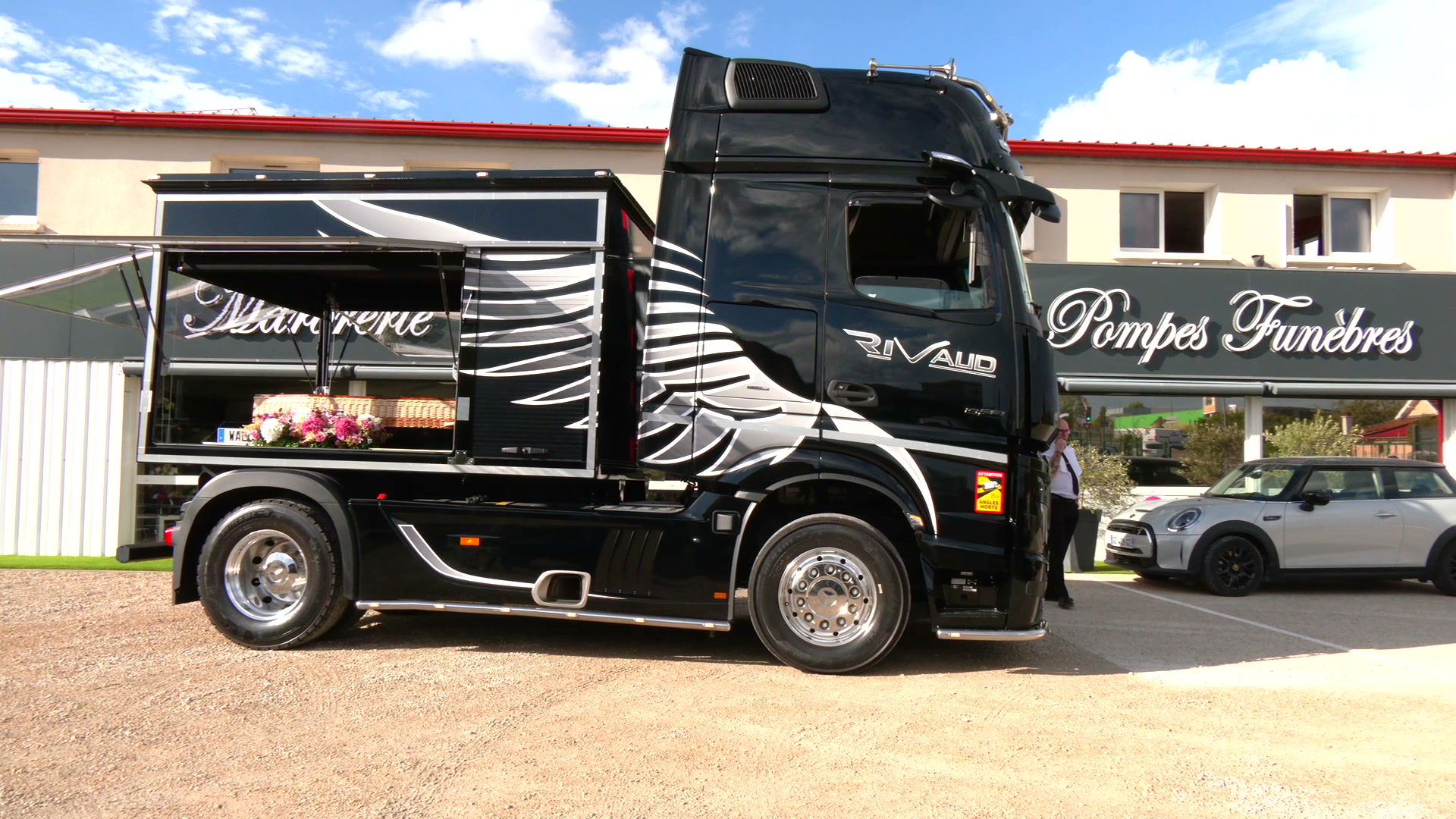 Camion corbillard noir aux motifs graphiques argent garé devant une enseigne Pompes Funèbres, cercueil fleuri visible dans la remorque ouverte