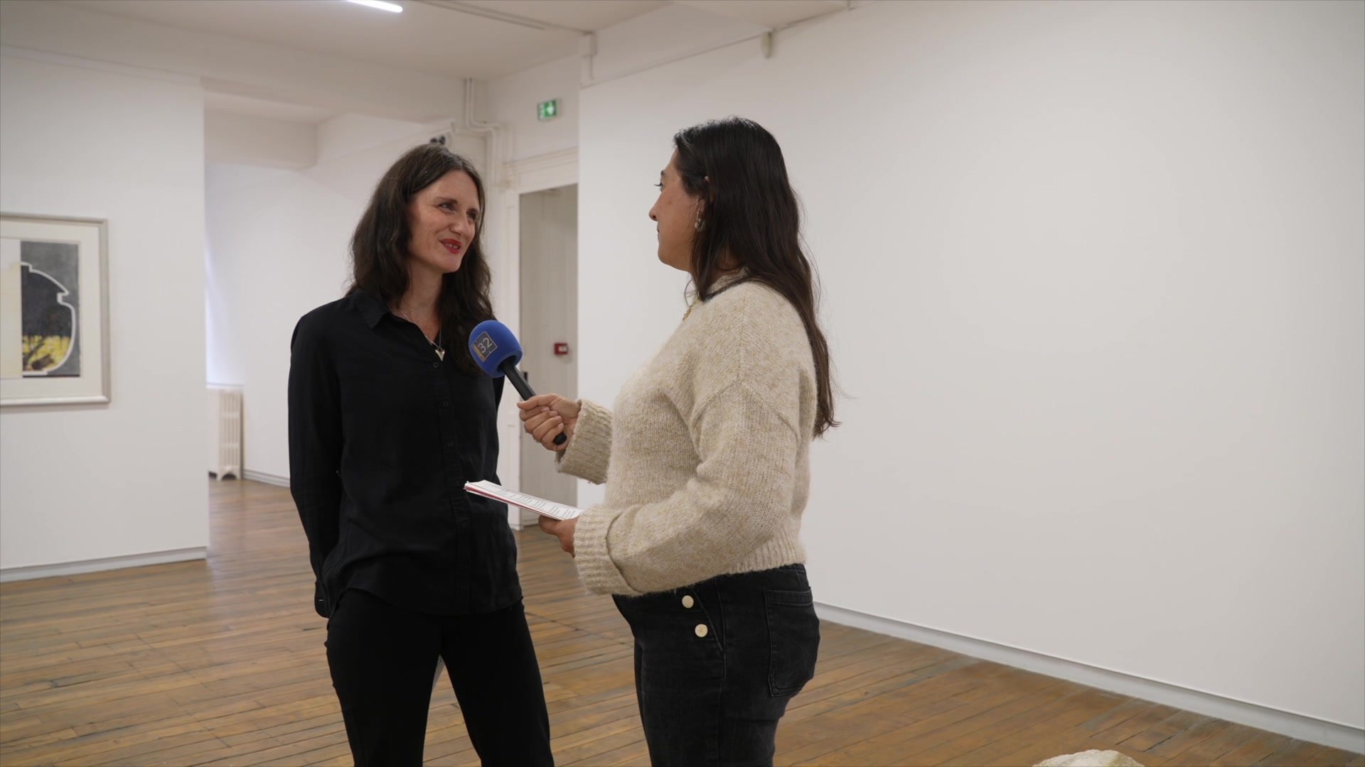 Deux femmes face à face dans une salle blanche aux parquet en bois, l'une tenant un micro Canal 32 bleu, l'autre les bras croisés, lors d'une interview à Passages