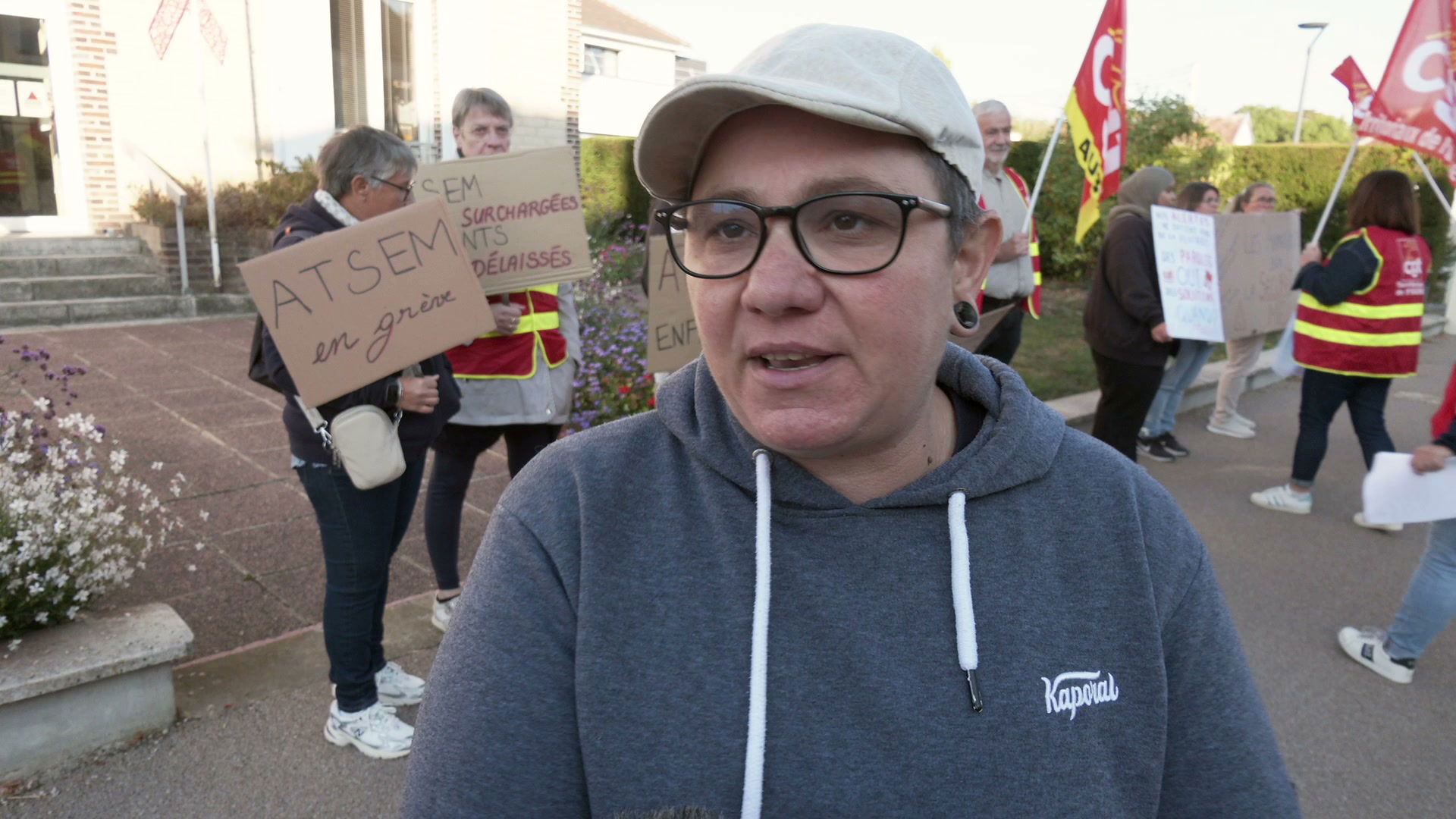Gréviste en sweat gris Kaporal et casquette beige au premier plan, manifestants avec pancartes ATSEM et drapeaux CGT derrière