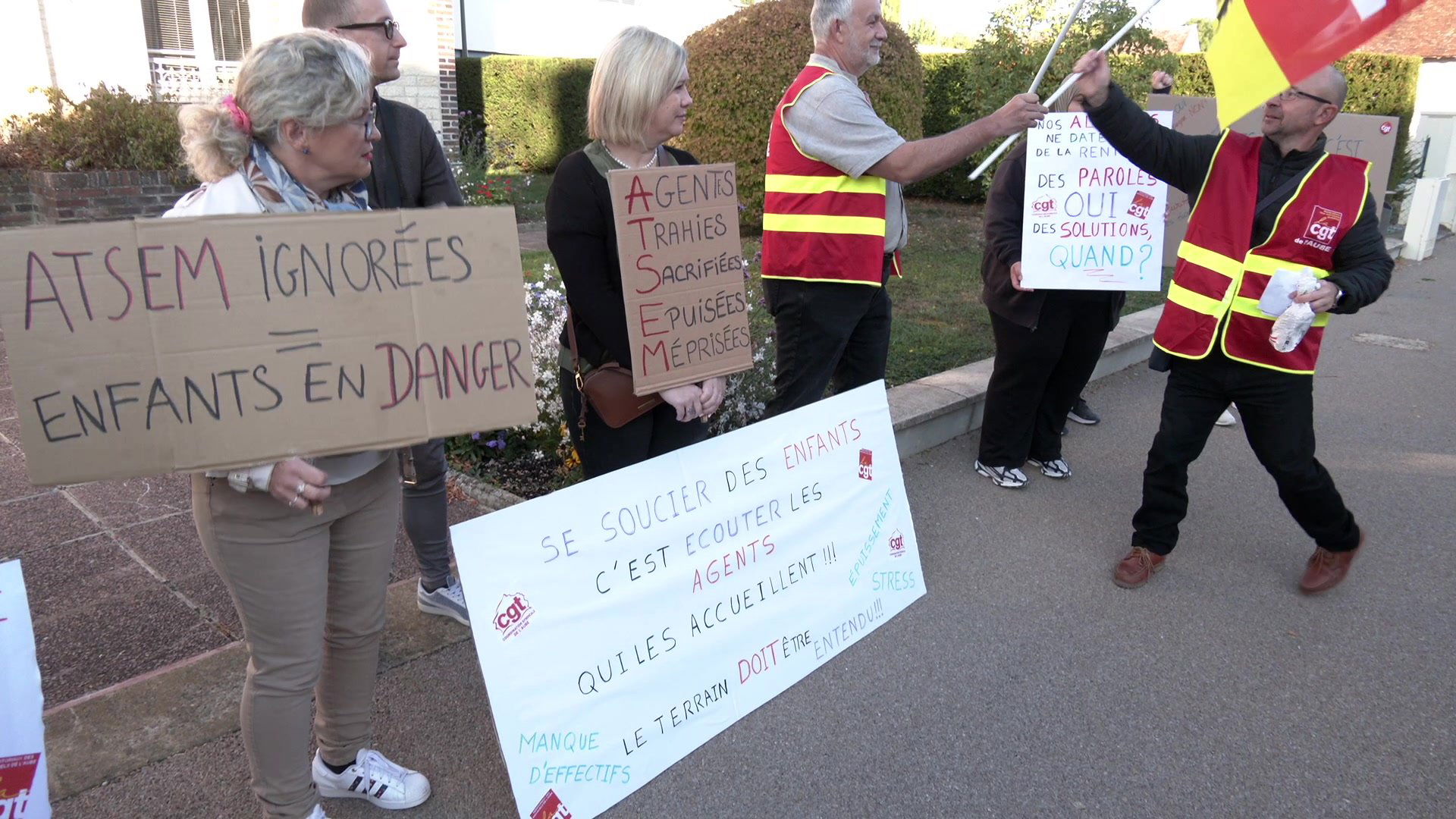 Gréviste en sweat gris au premier plan, pancarte ATSEM en grève et drapeaux CGT derrière elle devant la mairie de Bréviandes