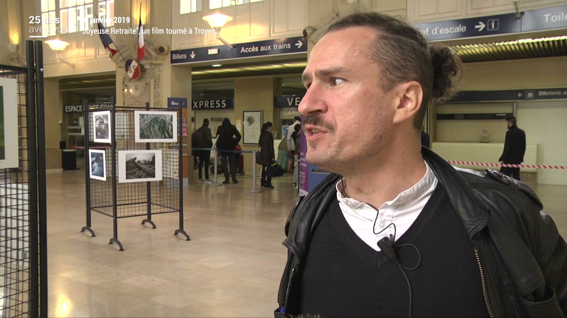 Homme interviewé dans le hall de la gare de Troyes, panneaux photos en arrière-plan