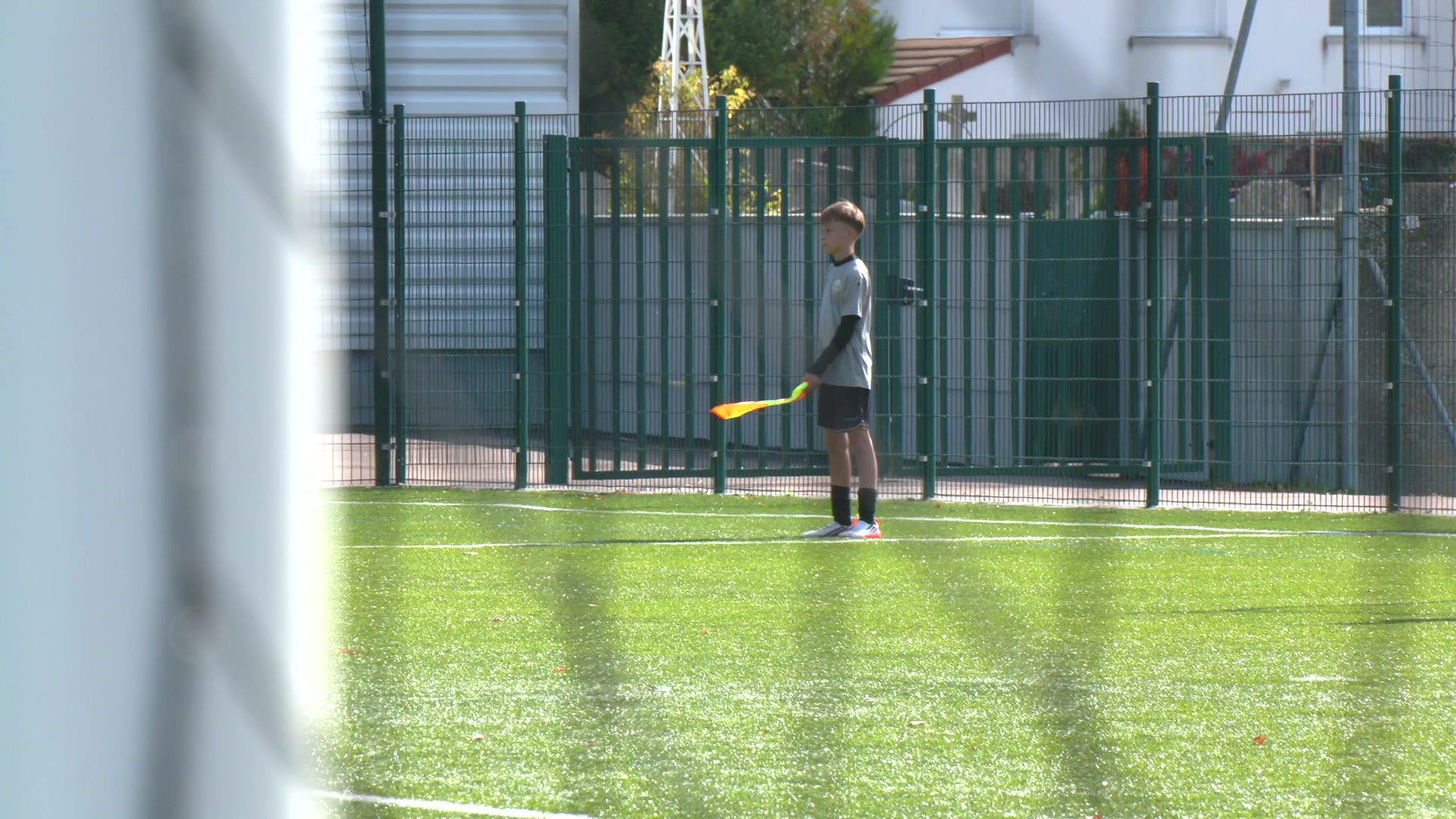 Jeune joueur U13 tenant un drapeau de touche orange et jaune sur un terrain synthétique vert