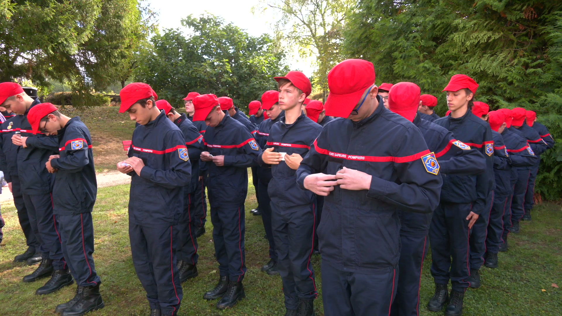 Jeunes sapeurs-pompiers en tenue bleu marine et casquette rouge alignés dans un jardin arboré lors de la remise de galons