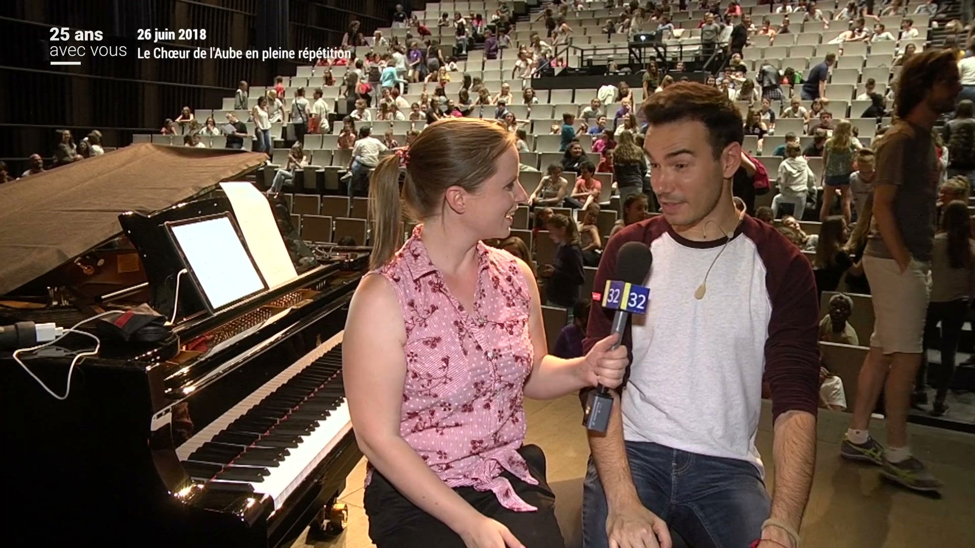 Journaliste Canal 32 interviewant une femme devant un piano à queue, salle de spectacle remplie de collégiens en