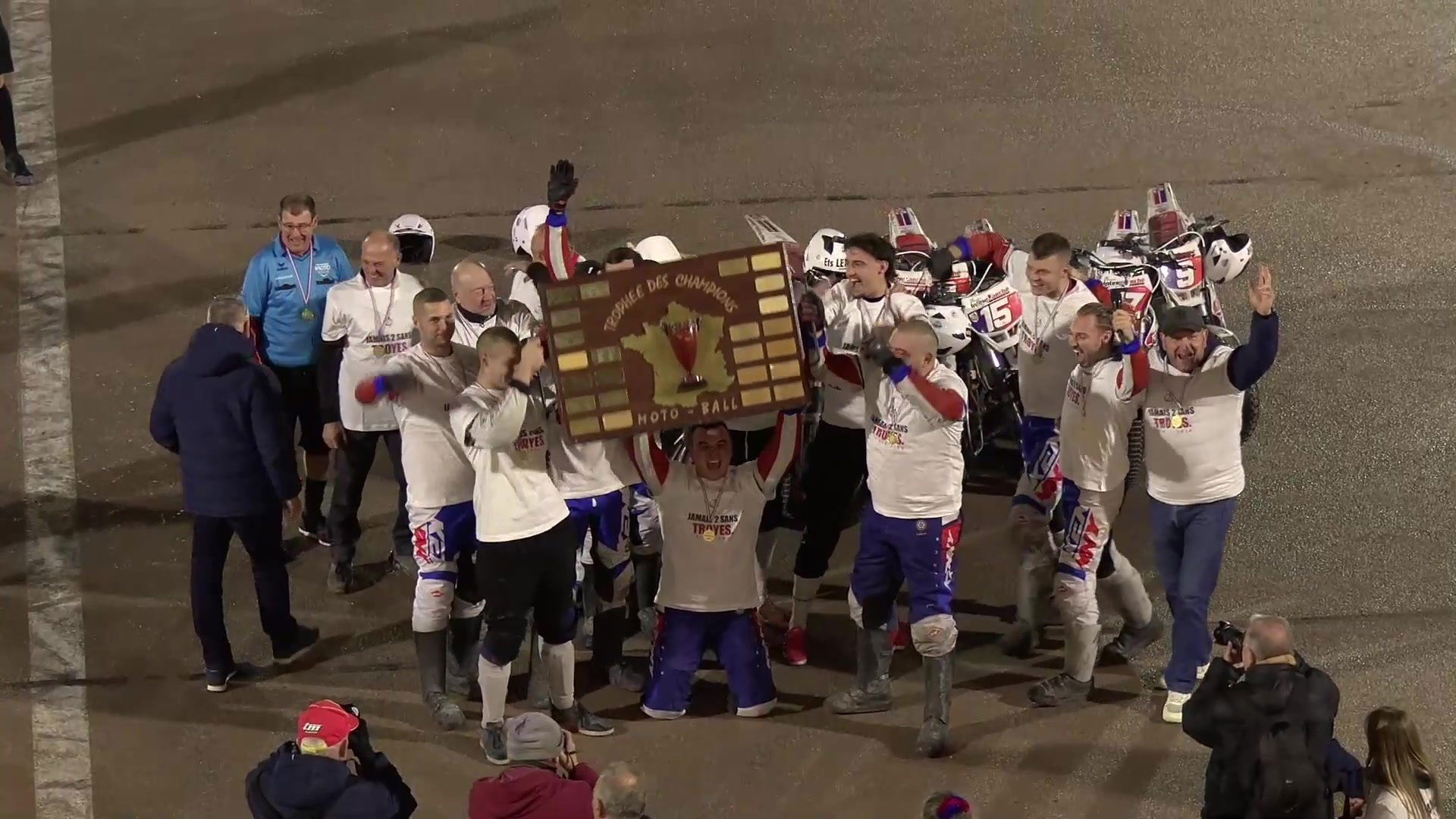 Les joueurs du SUMA en t-shirt blanc brandissent le bouclier du Trophée des champions Moto-Ball la nuit sur la piste du stade Gaston-Arbouin
