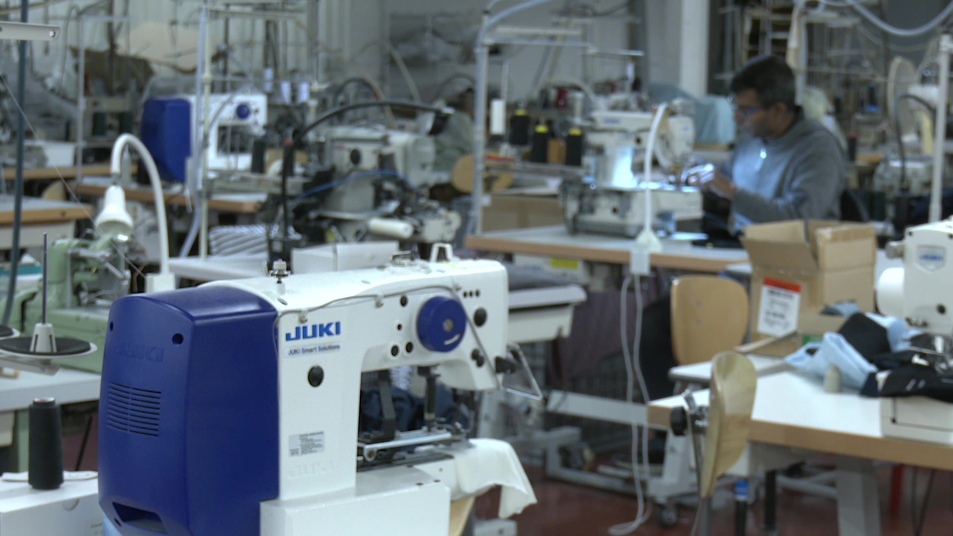 Machines à coudre Juki sur des tables de travail dans un atelier de confection industriel, avec un ouvrier en pull gris travaillant en arrière-plan