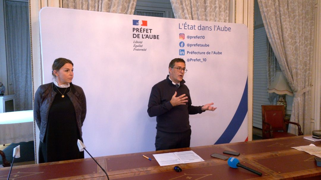 Mains d'un homme croisées devant lui, vêtu d'une veste sombre, fond beige clair légèrement flou