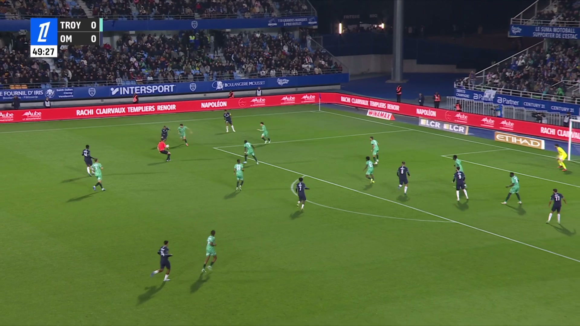Vue aérienne du Stade de l'Aube, score 0-0 à la 49e minute, joueurs en maillot bleu marine et vert sur la pelouse illuminée