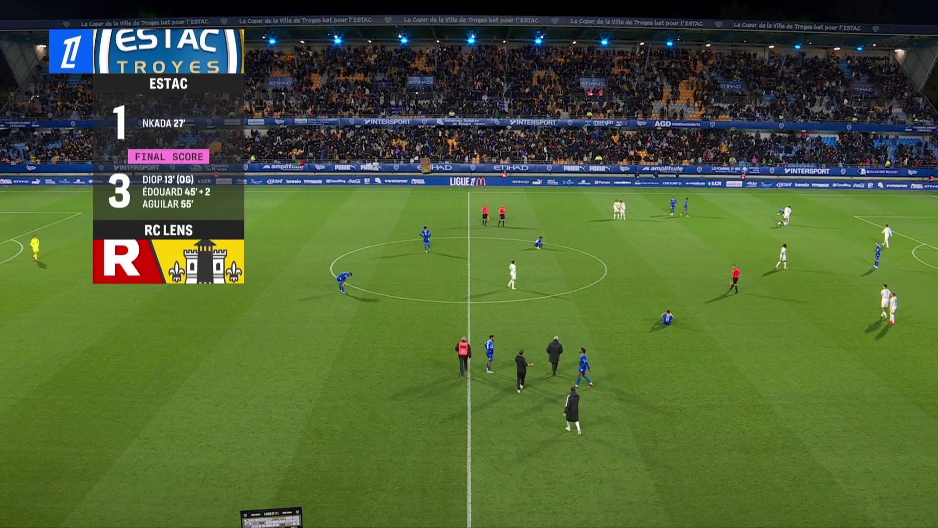 Vue aérienne du stade de l'Aube lors de Estac-Lens, score 1-1 affiché en première mi-temps, joueurs en maillot bleu et blanc sur la pelouse devant des tribunes pleines