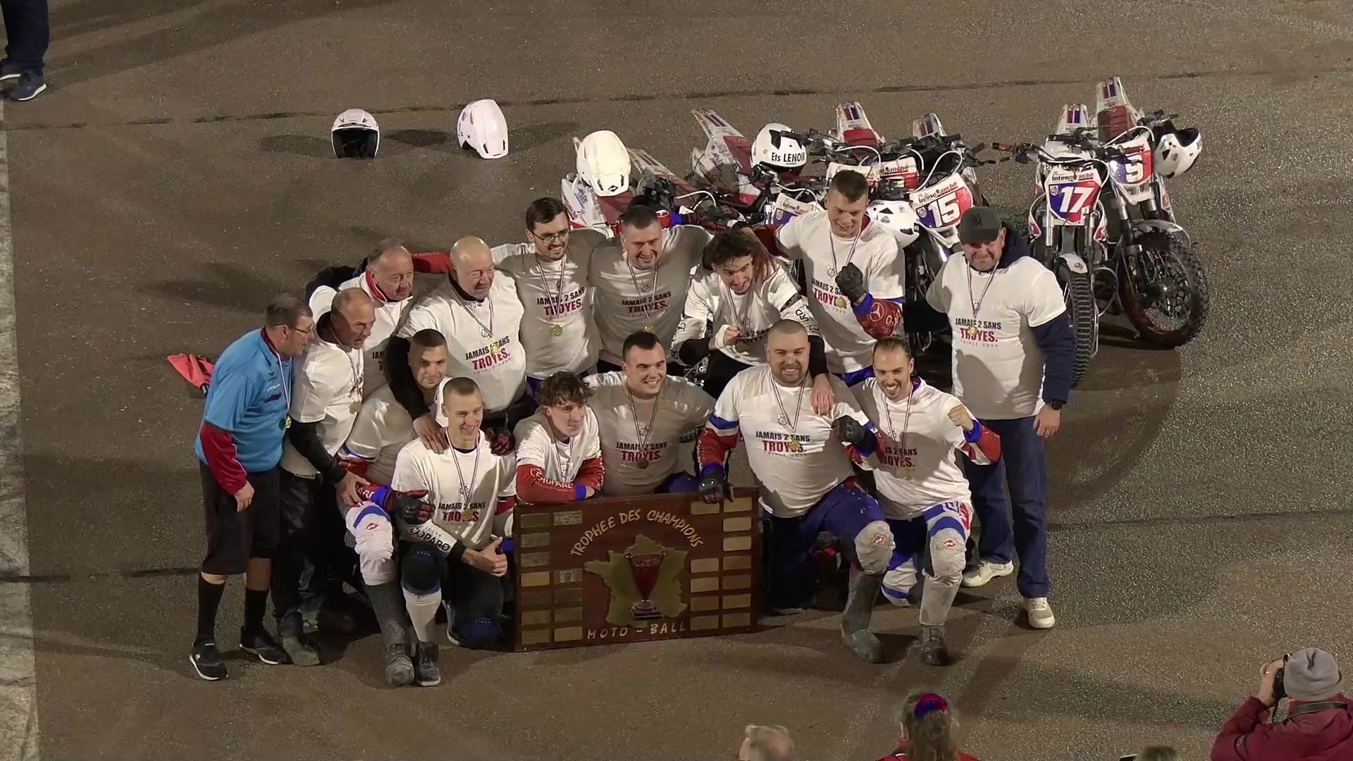 Vue aérienne du terrain de motoball avec deux équipes alignées face à face avec leurs motos, encadrées par des joueurs debout, panneau bleu trophée des champions affiché